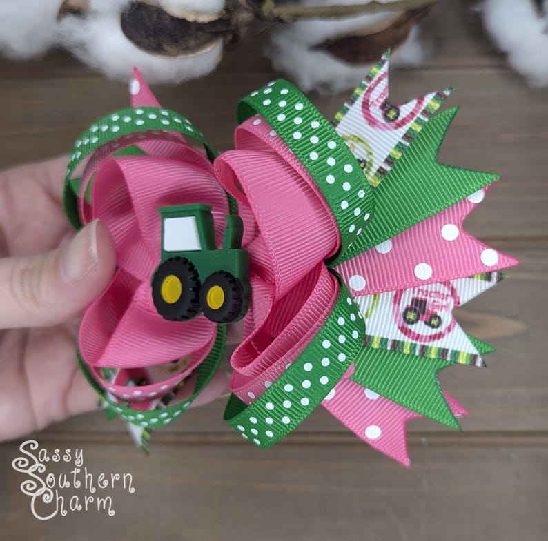 Puede incluir: Un lazo para el pelo rosa y verde con lunares blancos y un amuleto de tractor verde. El lazo est&aacute; hecho de cinta y est&aacute; atado en un nudo grande y esponjoso.