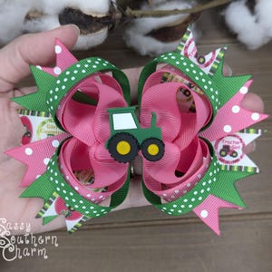 Puede incluir: Un lazo para el pelo rosa y verde con un amuleto de tractor verde. El lazo tiene lunares blancos y un estampado de ni&ntilde;a de tractor verde y blanco.