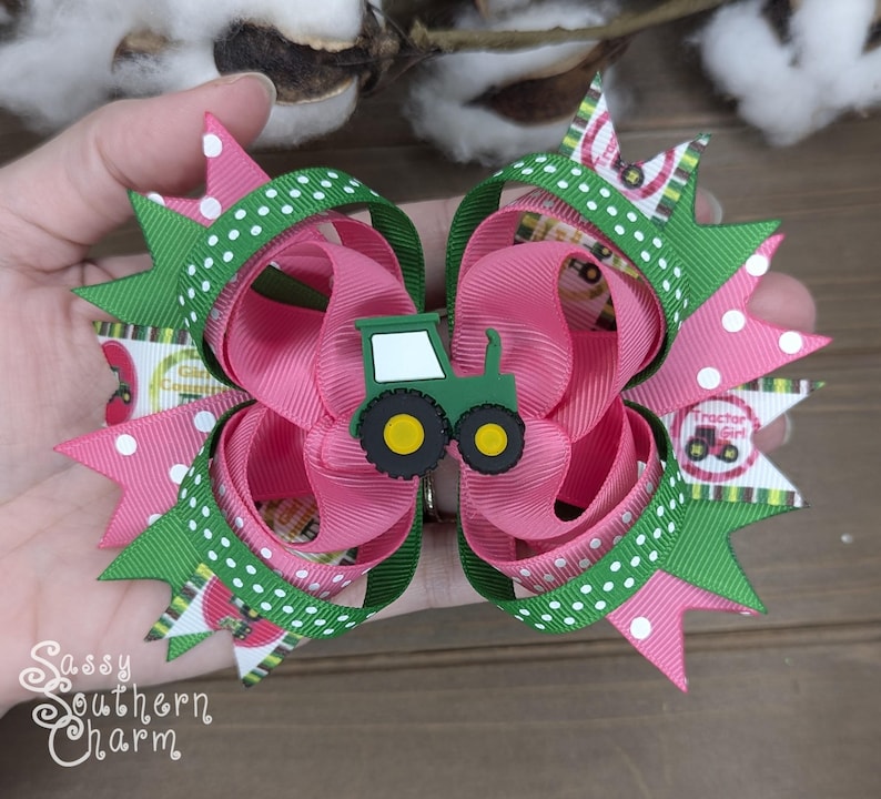 Puede incluir: Un lazo para el pelo rosa y verde con un amuleto de tractor verde. El lazo tiene lunares blancos y un estampado de ni&ntilde;a de tractor verde y blanco.