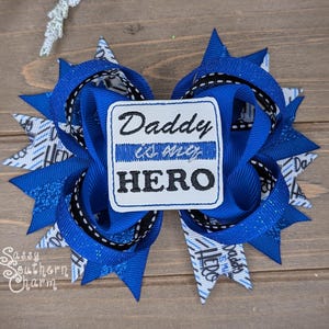 May include: A handmade blue hair bow with a white patch that reads "Daddy is my HERO" in black lettering. The bow features layers of blue ribbon, black and white patterned ribbon, and glitter accents. A decorative hair accessory.