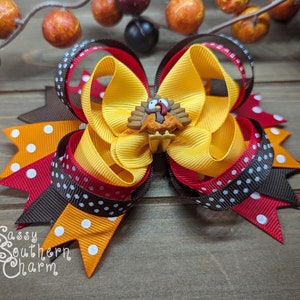 K&ouml;nnte beinhalten: Eine handgefertigte Haarschleife mit Thanksgiving-Motiv. Die Schleife hat gelbe, orangefarbene, rote und braune B&auml;nder, einige mit wei&szlig;en Punkten. Eine kleine Truthahn-Verzierung ist auf der Schleife zentriert. Der Text "Sassy Southern Charm" ist sichtbar.