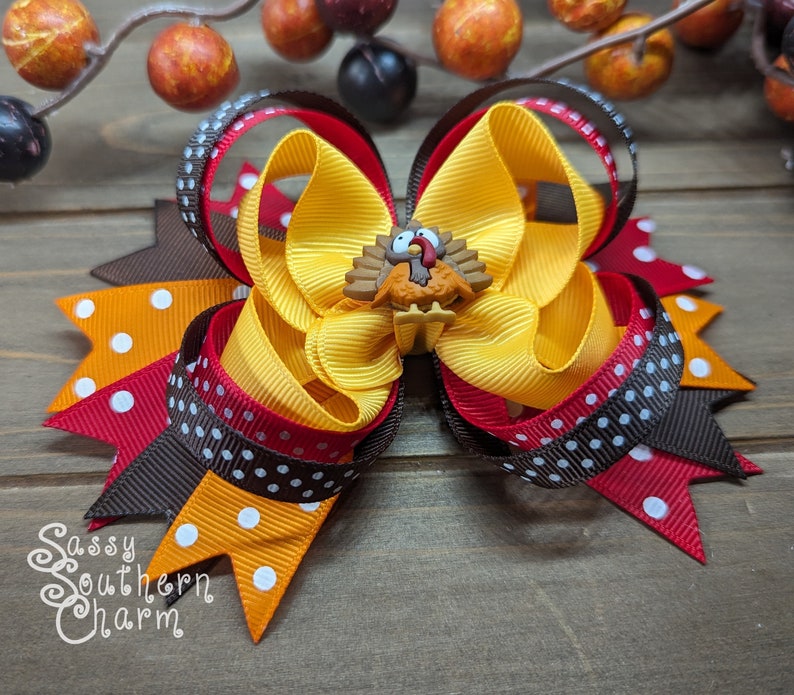K&ouml;nnte beinhalten: Eine handgefertigte Haarschleife mit Thanksgiving-Motiv. Die Schleife hat gelbe, orangefarbene, rote und braune B&auml;nder, einige mit wei&szlig;en Punkten. Eine kleine Truthahn-Verzierung ist auf der Schleife zentriert. Der Text "Sassy Southern Charm" ist sichtbar.