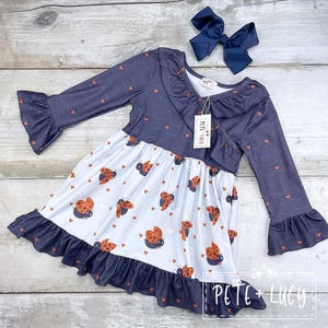 May include: A navy blue and white dress with a ladybug print and ruffle trim. The dress has long sleeves with ruffles at the cuffs and a crossover bodice. The dress is labeled "Pete + Lucy".