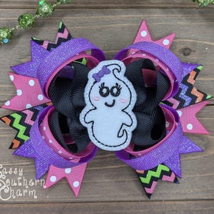 Peut inclure: Un n&oelig;ud pour cheveux violet, rose et noir avec une applique de fant&ocirc;me blanche. Le fant&ocirc;me a un n&oelig;ud violet et un visage souriant.