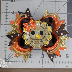May include: A brown, orange, and gold glitter hair bow with a felt turkey applique. The turkey is brown, gold, and yellow with white eyes and a red beak.