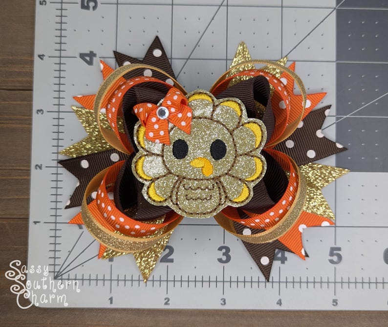 May include: A brown, orange, and gold glitter hair bow with a felt turkey applique. The turkey is brown, gold, and yellow with white eyes and a red beak.