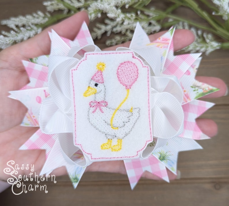 Puede incluir: Un lazo para el cabello hecho a mano con un dise&ntilde;o blanco y rosa. El lazo tiene un parche bordado central de un ganso con un gorro de fiesta y un globo rosa. El texto "Sassy Southern Charm" es visible.
