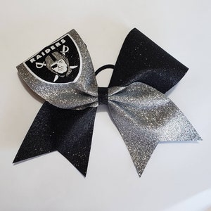 May include: A black and silver glitter cheer bow with the Las Vegas Raiders logo on one side.