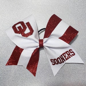 May include: A white and red glitter cheerleading bow. The bow features the letters "OU" in red glitter on one side and the word "SOONERS" in red glitter on the other side. The bow is tied with a black elastic band.
