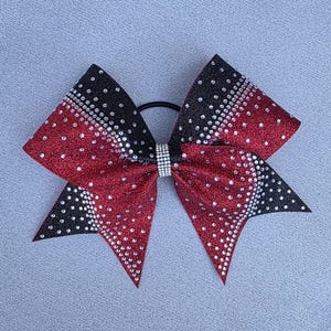 May include: A red and black glitter cheerleading bow with rhinestone accents. The bow is attached to a black elastic band and has a rhinestone center piece. The bow is a vibrant accessory.