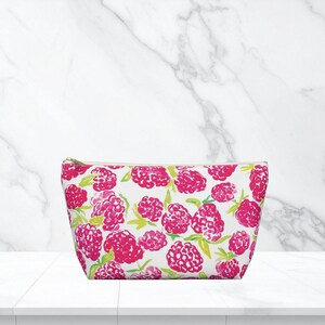 Puede incluir: Un neceser blanco con un vibrante estampado de frambuesas rosas y hojas verdes. La bolsa tiene una cremallera dorada y está diseñada para guardar maquillaje o artículos de tocador. La bolsa está hecha de un material duradero.