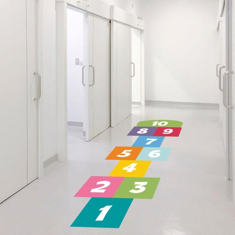 K&ouml;nnte beinhalten: Ein farbenfrohes H&uuml;pfspiel ist auf einem wei&szlig;en Boden in einem Flur aufgemalt. Die Quadrate sind mit den Zahlen 1 bis 10 versehen und haben verschiedene Farben, darunter T&uuml;rkis, Rosa, Gelb, Orange und Lila. Wei&szlig;e T&uuml;ren sind im Hintergrund zu sehen.