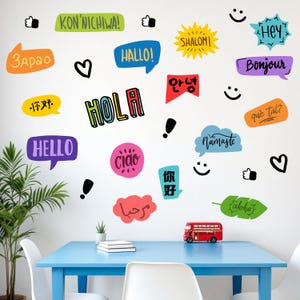 May include: A white wall decorated with colorful speech bubbles, hearts, thumbs-up symbols, and smiley faces. The speech bubbles contain greetings in various languages, including "Hello," "Hola," and "Bonjour." A red double-decker bus model sits on a blue table.