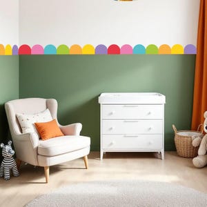 Könnte beinhalten: Ein Kinderzimmer mit einer weißen Wickelkommode, einem hellrosa Sessel und einer farbenfrohen Halbkreisbordüre. Ein orangefarbenes Kissen liegt auf dem Stuhl. Ein Stoffzebra und ein Teddybär befinden sich ebenfalls im Raum. Die Wände sind grün und weiß gestrichen.
