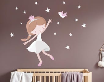 May include: A wall decal featuring a cartoon princess with a pink crown, white dress, and pink shoes, reaching for a pink bird. White star decals are scattered around the princess on a brown background. A wooden crib is in the foreground.
