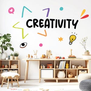 Może przedstawiać: Biała ściana z napisem "CREATIVITY" dużymi czarnymi literami, otoczona kolorowymi bazgrołami. Drewniane biurko i regał znajdują się na pierwszym planie, wypełnione elementami dekoracyjnymi i roślinami. Mały stołek z futrzanym siedziskiem stoi przed regałem.