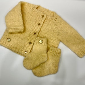 May include: A yellow knitted baby cardigan with button fastenings and matching socks. The cardigan and socks have small gold buttons with the words "Mommy & Me" on them.