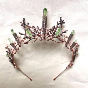 Può includere: Una corona artigianale color rame con accenti di cristalli verdi e delicati dettagli di foglie e rami. Il design della corona incorpora più cristalli verdi. La corona misura circa 18 cm di larghezza.
