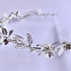May include: A silver-toned floral crown with delicate leaves and flower details. The crown is designed with a curved shape, and the leaves and flowers are intricately crafted. The crown is likely for a special occasion, such as a wedding or a formal event.