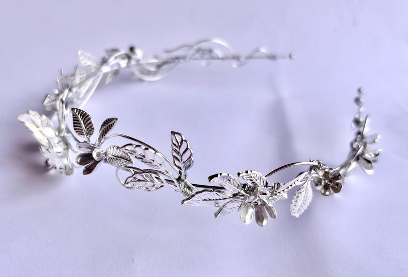 May include: A silver-toned floral crown with delicate leaves and flower details. The crown is designed with a curved shape, and the leaves and flowers are intricately crafted. The crown is likely for a special occasion, such as a wedding or a formal event.