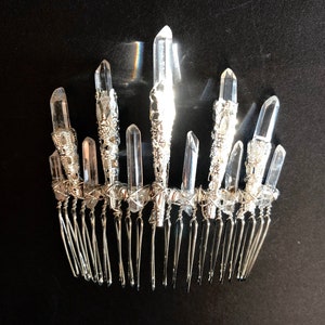 May include: A silver-toned hair comb adorned with clear quartz crystals. The crystals are of varying sizes and are set in ornate silver settings. The comb's teeth are long and slender, and the overall design is elegant and eye-catching.