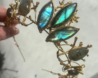 Op de afbeelding: Een goudkleurige metalen hoofdband met een takachtige structuur, versierd met meerdere iriserende labradoriet edelstenen. De stenen zijn in verschillende vormen geslepen, waaronder marquise en druppelvorm, en vertonen blauwe en groene lichtreflecties.