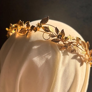 May include: A gold-colored floral crown with detailed leaves and small flowers. The crown is placed on a white surface, with a dark background. The crown's design features intricate details, suggesting a handcrafted or vintage style, suitable for special occasions.