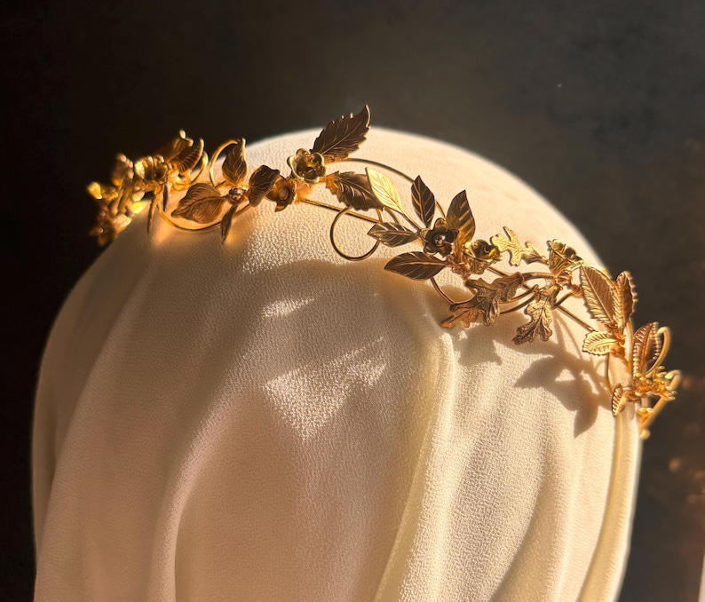 May include: A gold-colored floral crown with detailed leaves and small flowers. The crown is placed on a white surface, with a dark background. The crown's design features intricate details, suggesting a handcrafted or vintage style, suitable for special occasions.