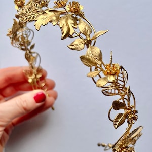 May include: A gold-colored floral crown with intricate leaf and flower details. The crown features a delicate, curved design with various sizes of leaves and flowers, creating an elegant and ornate appearance. The metal is a bright, shiny gold.