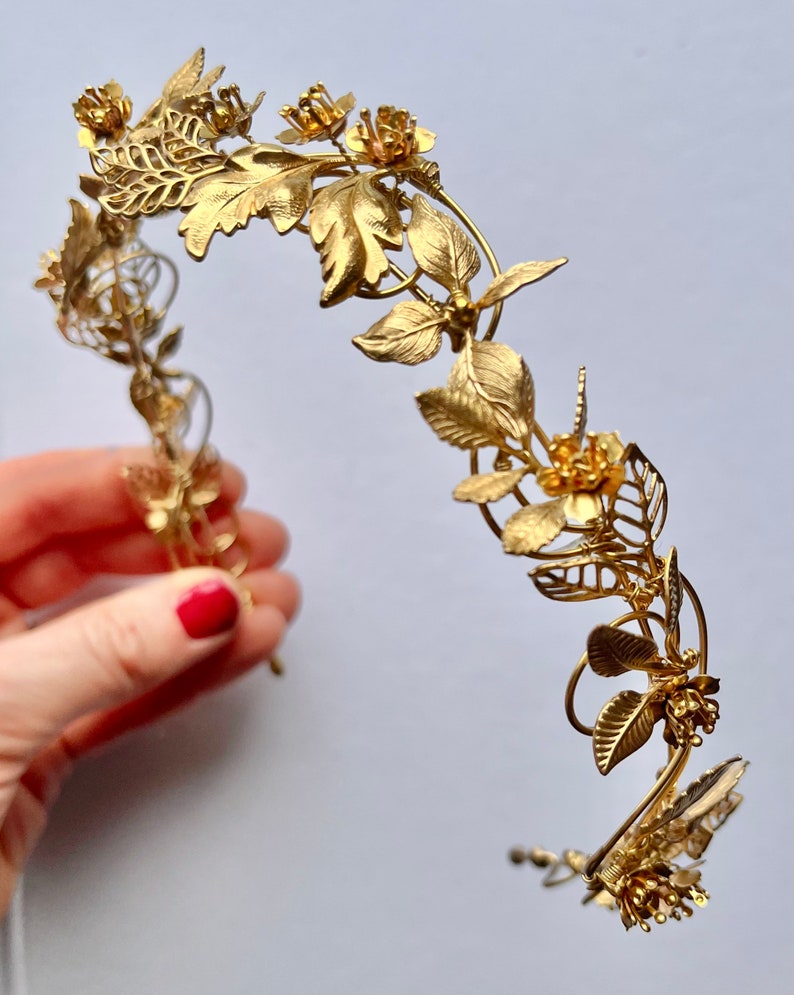 May include: A gold-colored floral crown with intricate leaf and flower details. The crown features a delicate, curved design with various sizes of leaves and flowers, creating an elegant and ornate appearance. The metal is a bright, shiny gold.