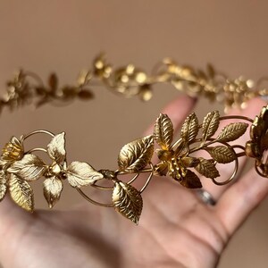 May include: A gold-toned metal crown featuring a delicate, continuous vine design. The headpiece is adorned with intricate, textured leaves and small, detailed flower blossoms arranged symmetrically along a thin, flexible wire frame.