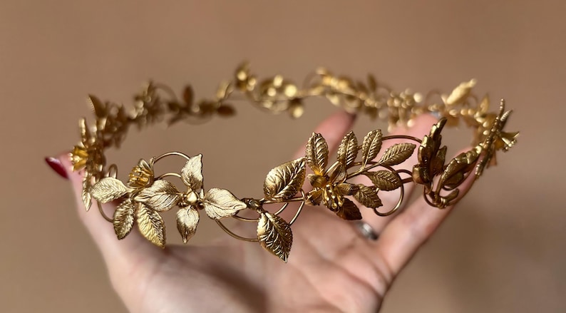 May include: A gold-toned metal crown featuring a delicate, continuous vine design. The headpiece is adorned with intricate, textured leaves and small, detailed flower blossoms arranged symmetrically along a thin, flexible wire frame.