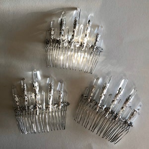 May include: Three silver hair combs with clear crystal accents. Each comb features a row of tines and is adorned with multiple crystal formations. The crystals are clear and faceted, catching the light. The combs are arranged on a light-colored surface.