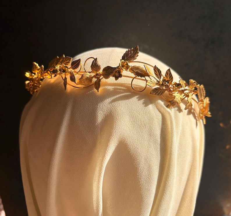 May include: A gold-colored floral crown with delicate leaves and flowers. The crown is made of thin, flexible metal and is designed to be worn on the head. The leaves and flowers are intricately detailed, giving the crown a natural and elegant appearance.