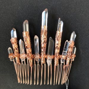 May include: A copper-colored hair comb featuring clear quartz crystal points. The crystals are secured with ornate copper-colored metalwork. The comb's teeth are made of copper-colored wire, creating a unique and elegant hair accessory.