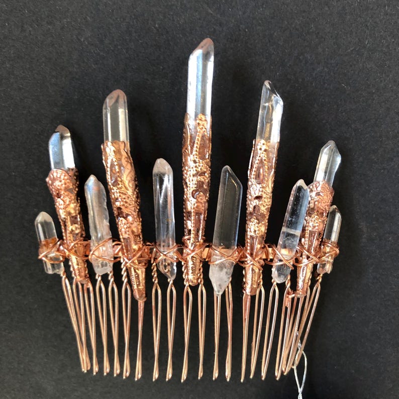 May include: A copper-colored hair comb featuring clear quartz crystal points. The crystals are secured with ornate copper-colored metalwork. The comb's teeth are made of copper-colored wire, creating a unique and elegant hair accessory.