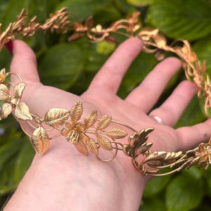 May include: A circular headpiece crafted from metallic gold-toned wire, featuring intricate, detailed leaves and small flower blossoms arranged in a continuous vine-like pattern that forms a delicate, open-structured crown design.