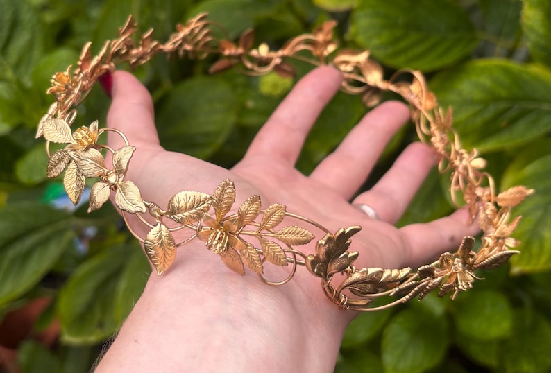 May include: A circular headpiece crafted from metallic gold-toned wire, featuring intricate, detailed leaves and small flower blossoms arranged in a continuous vine-like pattern that forms a delicate, open-structured crown design.