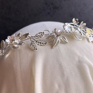 May include: A silver-toned floral crown featuring delicate flowers and leaves. The crown is designed with intricate details, showcasing a botanical theme. The metal crown is displayed on a white surface against a dark background.