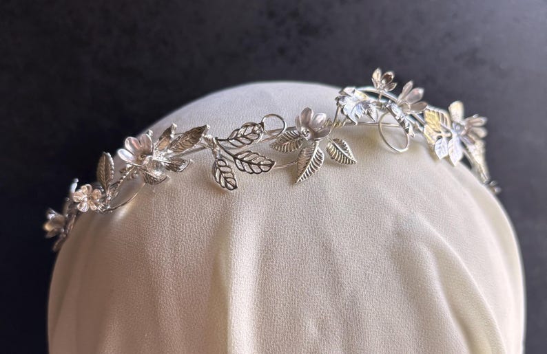 May include: A silver-toned floral crown featuring delicate flowers and leaves. The crown is designed with intricate details, showcasing a botanical theme. The metal crown is displayed on a white surface against a dark background.