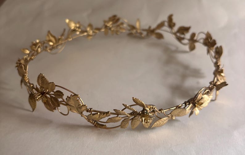 May include: A gold-toned metal crown featuring a delicate vine-like structure adorned with small, textured leaves and tiny flower blossoms. The circular headpiece is constructed from thin, flexible wire, creating an intricate botanical design.