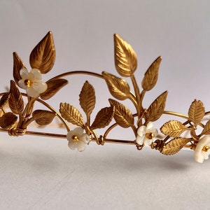 Könnte beinhalten: Ein goldener Blätter- und weißer Blumenkranz. Die Krone ist aus Metall und hat ein zartes Design.