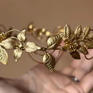 May include: A gold-toned metal crown featuring a delicate arrangement of textured leaves and small flowers. The headpiece is constructed from thin wire branches that form a circular band, showcasing intricate botanical details throughout the design.