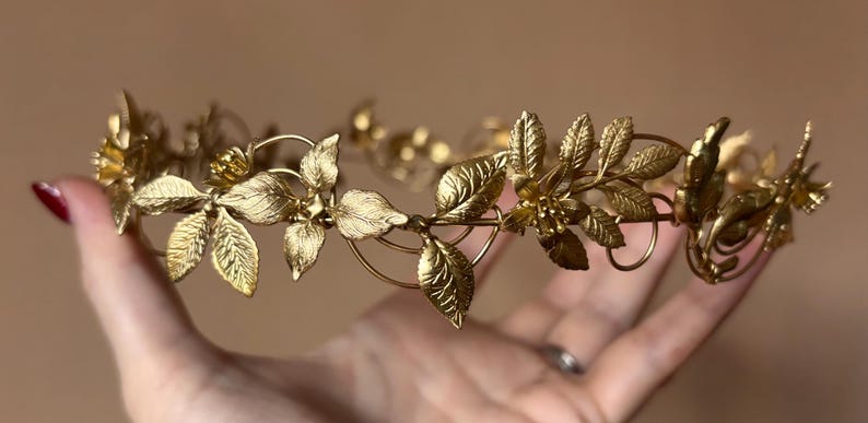 May include: A gold-toned metal crown featuring a delicate arrangement of textured leaves and small flowers. The headpiece is constructed from thin wire branches that form a circular band, showcasing intricate botanical details throughout the design.