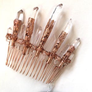 May include: A copper-colored hair comb adorned with clear quartz crystals. The crystals are set in ornate, filigree-style metalwork. The comb's tines are thin and evenly spaced, creating a delicate and elegant accessory.