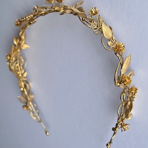 May include: A gold-colored floral crown with delicate leaves and flowers. The headpiece has a curved design with intricate details, perfect for weddings or special occasions. The crown is made of a shiny, metallic material.