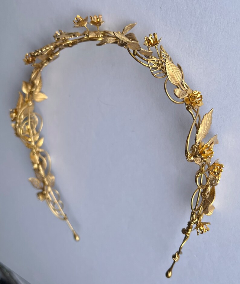May include: A gold-colored floral crown with delicate leaves and flowers. The headpiece has a curved design with intricate details, perfect for weddings or special occasions. The crown is made of a shiny, metallic material.
