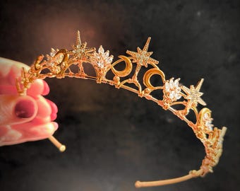 Op de afbeelding: Goudkleurige tiara met ster- en maanmotieven, versierd met heldere kristallen. De delicate, sierlijke structuur van de tiara maakt het een ideaal accessoire voor speciale gelegenheden of thema-evenementen.