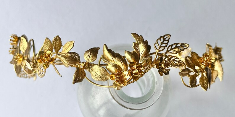 May include: A gold-colored floral crown with detailed leaves and flowers. The crown is made of metal and has a delicate, ornate design. The leaves and flowers are arranged around the band, creating a natural, elegant look.
