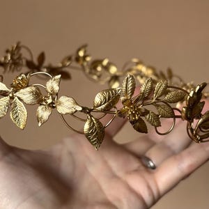 May include: A gold-toned metal crown featuring a delicate arrangement of textured leaves and small floral accents. The circular headpiece is constructed from thin, interconnected wire branches that form a continuous botanical pattern throughout.